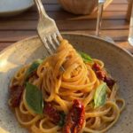 A close-up of a fork twirling creamy linguine with a piece of sun-dried tomato, with a glass of white wine in the background.