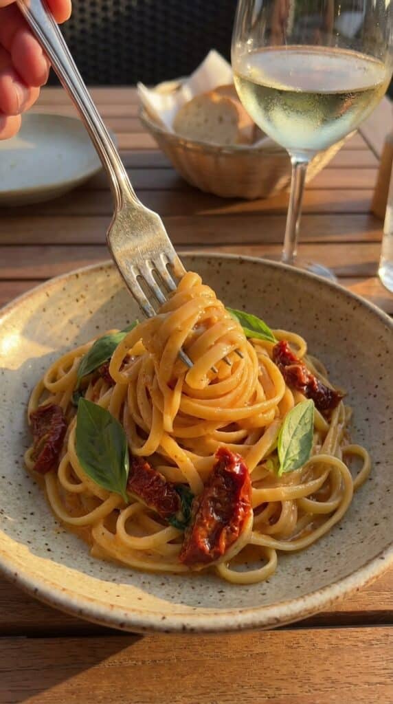 A close-up of a fork twirling creamy linguine with a piece of sun-dried tomato, with a glass of white wine in the background.