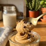 A close-up of a gold spoon lifting a thick, smooth dollop of peanut butter mousse dotted with chocolate chips and sea salt.