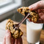 Two hands breaking a warm peanut butter oatmeal chocolate chip cookie in half, showing melting dark chocolate and a chewy center.