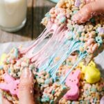 Two hands pulling apart a pastel pink and blue Rice Krispie treat, showing a long, gooey marshmallow stretch.