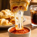 A close-up of a hand dipping a cheesy, flaky pizza crescent roll into marinara sauce, featuring a long cheese pull.