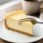 A close-up of a dense slice of New York cheesecake on a plate with a fork cutting into the tip, served with coffee.