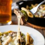 A close-up of a fork lifting a bite of cheesy steak pasta, creating a long stringy cheese pull.