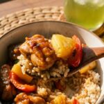 A close-up of a spoon lifting a bite of pineapple chicken and rice, featuring a chunk of pineapple, chicken, and bell pepper.