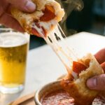 A close-up of a hand breaking apart a pizza roll with a long cheese pull, about to dip it into a bowl of marinara sauce.