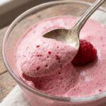 A close-up of a silver spoon lifting a scoop of airy, fluffy pink raspberry mousse from a glass.