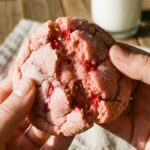 Two hands breaking a soft, pink raspberry sugar cookie in half, showing a chewy, doughy center with a glass of milk in the background.