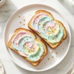 A close-up of a hand holding a piece of half-eaten rainbow unicorn toast with bright window light and a coffee mug in the background.