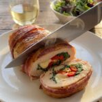 A close-up of a knife slicing a stuffed chicken breast in half, showing the melted cheese, spinach, and red pepper filling inside.