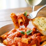 A close-up of a fork twirling rigatoni pasta coated in a thick, creamy red pepper sauce over a white bowl.