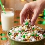 A close-up of a hand grabbing a handful of powdered sugar puppy chow with green M&Ms from a bowl.