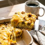 A close-up of a spatula lifting a cheesy square of breakfast casserole out of the pan, showing a cheese pull, with coffee in the background.