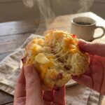 A close-up of hands pulling apart a warm ham and cheese scone, showing the steam and melted cheese inside, with butter melting on the crumb.