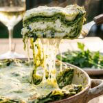 A close-up of a spatula lifting a slice of green spinach lasagna with a long stringy cheese pull, showing the distinct layers.