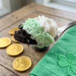 A close-up of a spoon lifting a scoop of green mint pudding, chocolate crust, and whipped cream out of a dessert cup.