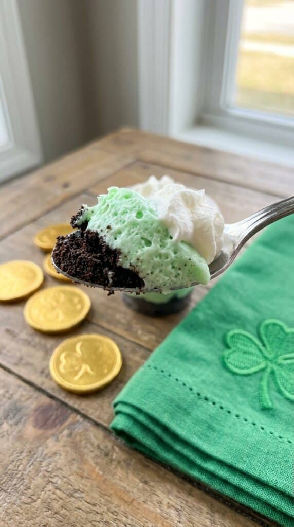 A close-up of a spoon lifting a scoop of green mint pudding, chocolate crust, and whipped cream out of a dessert cup.