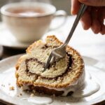 A close-up of a fork cutting into a slice of glazed snickerdoodle bundt cake, showing the dark cinnamon swirl inside.