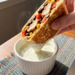 A close-up of a hand dipping a toasted Southwest chicken wrap half into a small bowl of creamy dressing.