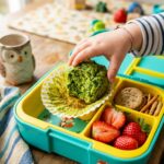 A kid's hand reaching for a bright green spinach muffin packed inside a colorful bento lunchbox with strawberries.