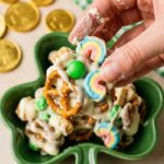 A close-up of a hand grabbing a handful of white chocolate coated St. Patrick's Day snack mix from a green bowl.