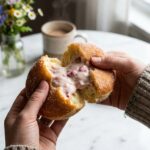 A close-up of hands pulling apart a sugar-coated donut, revealing a thick, creamy pink strawberry filling inside.