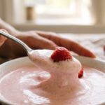 A close-up of a silver spoon lifting a thick, creamy dollop of pink strawberry mousse with a piece of fresh fruit.