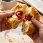 A close-up of hands pulling apart a strawberry danish, showing the flaky pastry layers and creamy fruit center, with coffee in the background.
