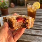 A close-up of a hand holding a strawberry lemon blondie with a bite taken out, showing a fudgy interior and baked strawberries.