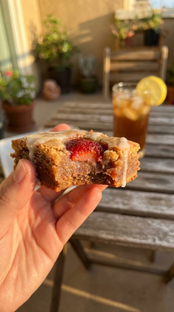 A close-up of a hand holding a strawberry lemon blondie with a bite taken out, showing a fudgy interior and baked strawberries.