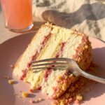 A close-up of a fork cutting through a tall slice of layered strawberry shortcake cheesecake, showing the fluffy cake, creamy center, and crumbly coating.