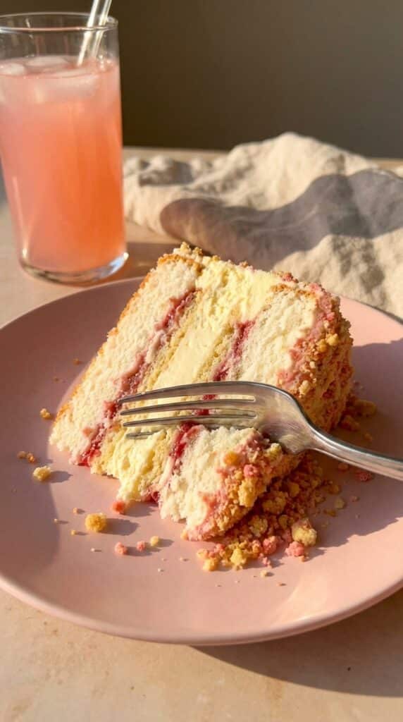A close-up of a fork cutting through a tall slice of layered strawberry shortcake cheesecake, showing the fluffy cake, creamy center, and crumbly coating.