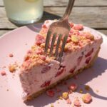A close-up of a fork cutting into a slice of strawberry freezer pie, showing the creamy pink filling and thick crunchy crumb layers on a pink plate.