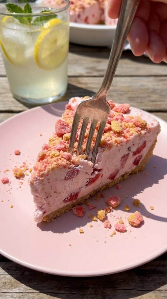 A close-up of a fork cutting into a slice of strawberry freezer pie, showing the creamy pink filling and thick crunchy crumb layers on a pink plate.