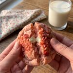 Two hands pulling apart a soft, pink strawberry sugar cookie showing a melted white chocolate chip inside, with a glass of milk in the background.