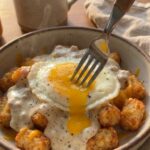 A close-up of a fork breaking a runny egg yolk over crispy tater tots and sausage gravy, with coffee in the background.