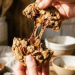 Two hands pulling apart a warm, thick cowboy cookie, showing a gooey melted chocolate pull and a soft oat and pecan center.