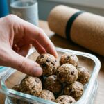A hand grabbing a chocolate chip protein ball out of a glass meal prep container, with a yoga mat blurred in the background.