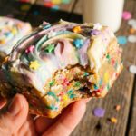 A close-up of a hand holding a half-eaten unicorn bar, showing the soft, chewy funfetti center and thick swirled frosting.