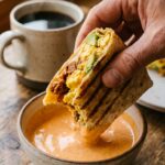 A hand holding a half-eaten, toasted breakfast burrito and dipping it into a bowl of spicy mayo with a coffee mug in the background.