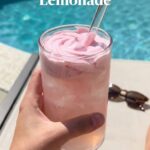 A close-up of a hand stirring thick pink whipped lemonade into iced water with a glass straw against a blurred swimming pool background.