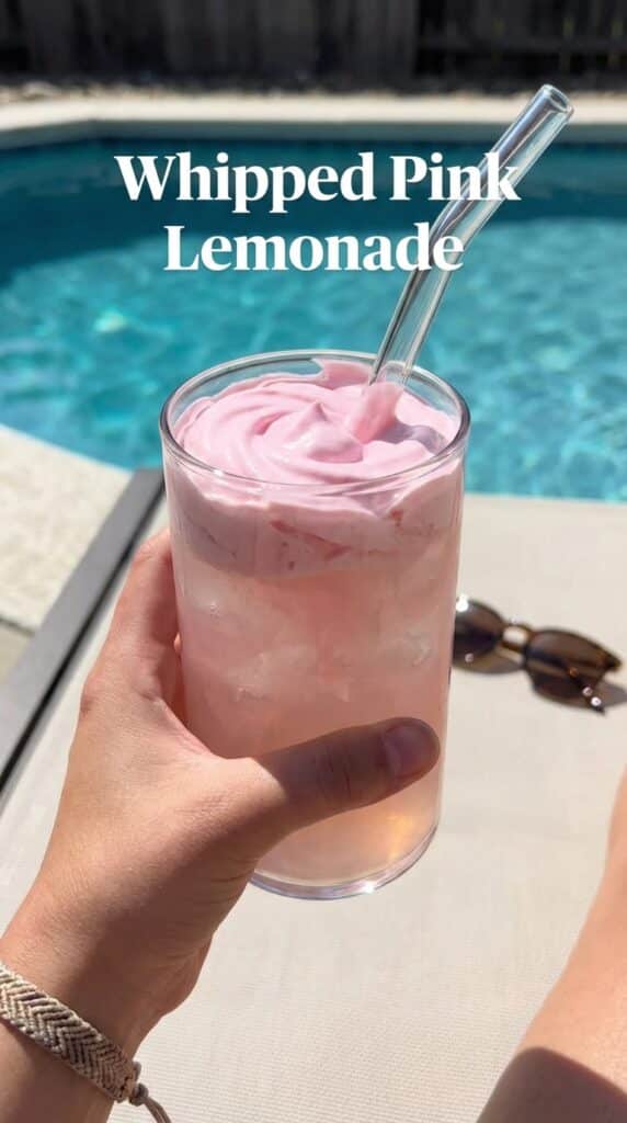 A close-up of a hand stirring thick pink whipped lemonade into iced water with a glass straw against a blurred swimming pool background.