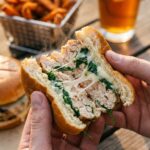 Two hands holding a halved chicken, spinach, and white cheddar burger, showing the juicy, cheesy interior with steam rising.
