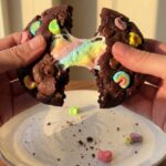 Two hands pulling apart a dark chocolate cookie, revealing a fudgy center and a stretching, gooey melted marshmallow.