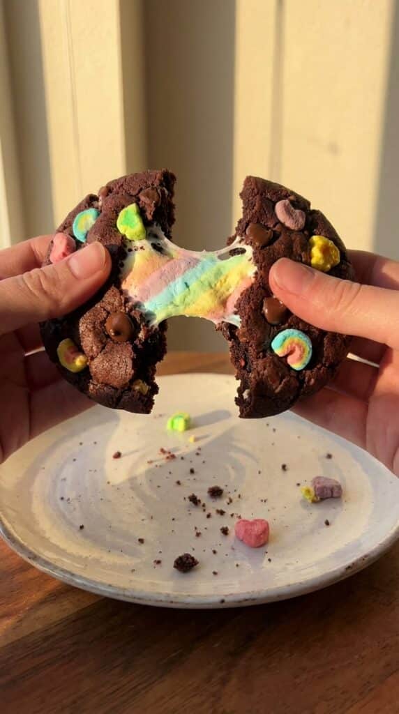 Two hands pulling apart a dark chocolate cookie, revealing a fudgy center and a stretching, gooey melted marshmallow.