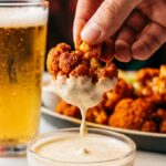 A close-up of a hand dipping a spicy cauliflower wing into ranch dressing.