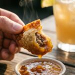 A close-up of a hand dipping a crispy wonton into mango sauce, showing the filling inside.