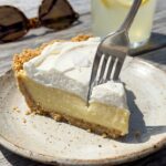 A close-up of a fork cutting into a slice of Atlantic Beach pie, showing the crumbly saltine crust, dense lemon filling, and whipped cream.