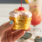 A tropical Bahama Mama cupcake with pink frosting, a cherry, pineapple wedge, and a paper umbrella on a bamboo tray by a pool.