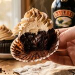 A close-up of a hand peeling the wrapper off a moist chocolate cupcake with a thick swirl of frosting.